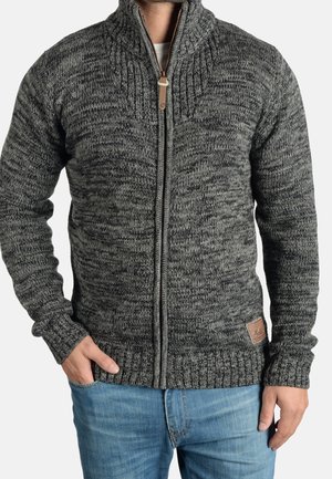 Homme portant un pull en maille gris zippé avec poignets et ourlet côtelés, associé à un jean bleu clair, debout, une main dans la poche.