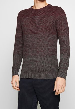 Homme portant un pull en maille texturée bordeaux et gris avec des poignets côtelés, assorti à un pantalon rayé foncé.