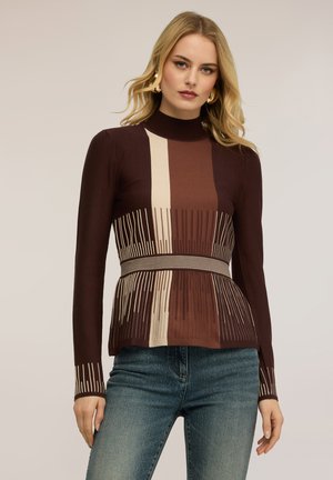 Donna bionda che indossa orecchini a cerchio dorati, maglia attillata a maniche lunghe con motivo marrone e beige, e jeans blu su sfondo neutro.