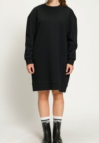 Robe sweat noire oversize avec des manches longues, des poignets côtelés et un col ras du cou, fabriquée à partir d'un tissu doux et texturé, portée avec des bottes zippées noires.
