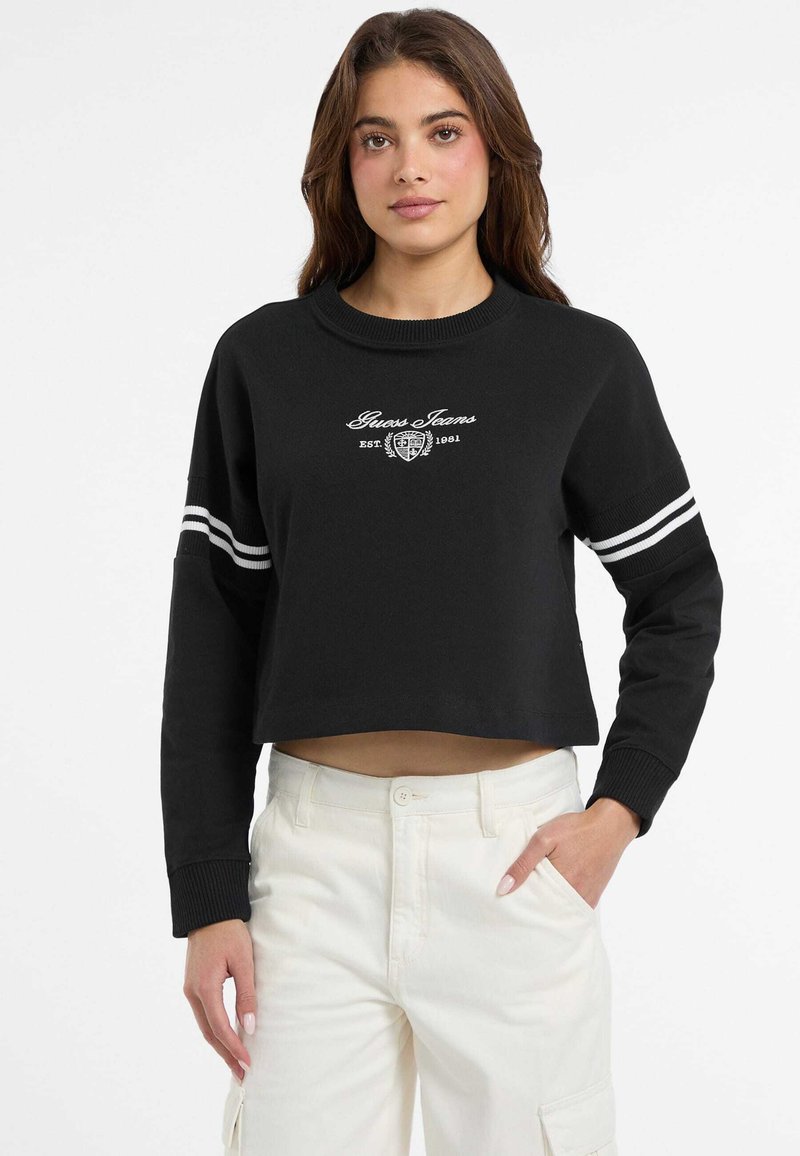 Felpa nera corta con logo ricamato bianco, caratterizzata da un collo rotondo a coste e polsini a righe. Abbinata a pantaloni bianchi.