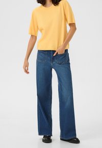 Top corto a maniche corte di colore giallo abbinato a jeans in denim blu a gamba larga, con tasche posteriori e vestibilità rilassata. Le calzature nere completano il look.