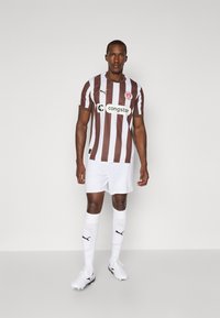 Brun og hvid stribet fodboldtrøje med logo, parret med hvide shorts og sokker, der har sorte accenter og fodtøj.