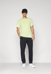 Hellgrünes Kurzarm-T-Shirt mit "Champion"-Logo, kombiniert mit dunklen Jogginghosen und weißen Sneakers, getragen mit einer schwarzen Cap.