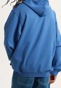 Personne aux cheveux foncés tressés portant un sweat à capuche bleu ample et un jean en denim bleu, vue de dos sur un fond uni.