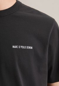 Čierne tričko s krátkym rukávom vyrobené z bavlny s lemovaným výstrihom. Na prednej strane má biele nápisy "MARC O’POLO DENIM". Hladká textúra.