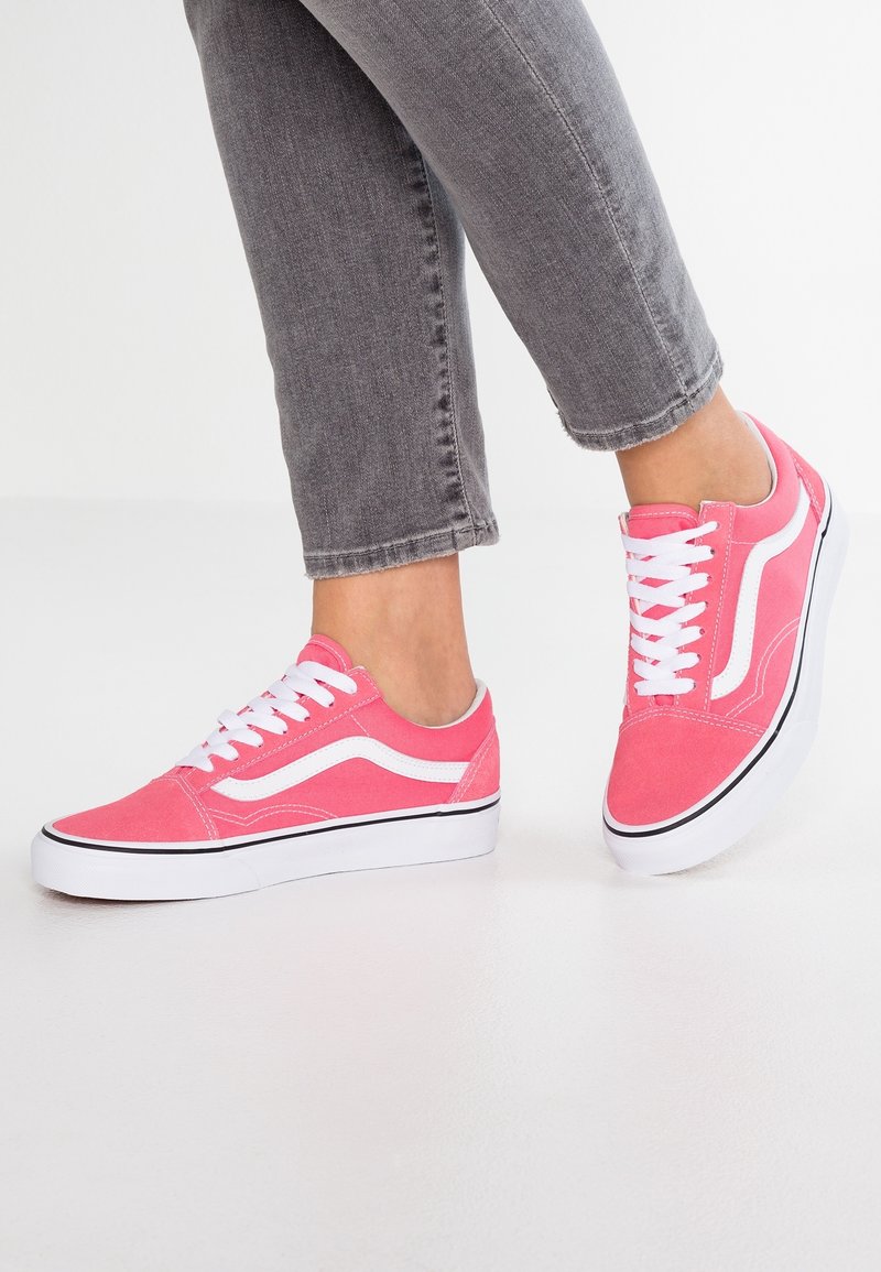 Pieds portant des baskets basses roses avec des lacets blancs et une bande ondulée, associées à un jean gris raccourci sur fond blanc.