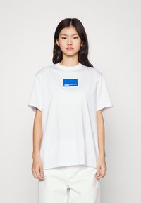 Hvid bomulds T-shirt med korte ærmer, der har et blåt rektangulært logo på brystet. Rund halsudskæring, afslappet pasform, glat tekstur.