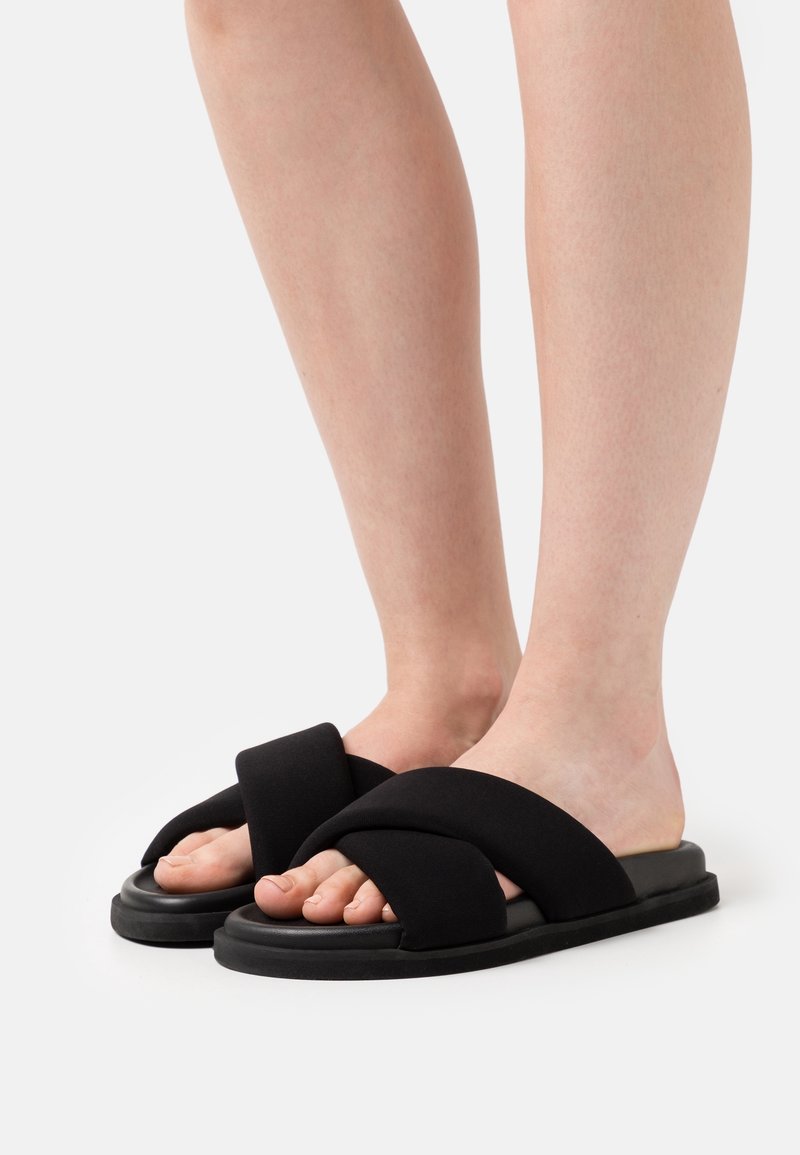 Pieds nus portant des sandales noires à enfiler avec des brides croisées épaisses et des semelles plates sur un fond clair.
