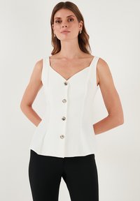 Fehér ujjatlan felső V-nyakkal, az elején öt fémből készült gombbal és fodros peplum szegéllyel. Simá szövet textúra.