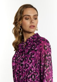 Paarse blouse met luipaardprint, voorzien van een gestrikt decolleté, doorzichtig materiaal en lichtgewicht stof. Aangevuld met grote, ronde oorbellen.
