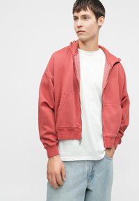 Σακάκι ροζ με φερμουάρ και κουκούλα με κορδόνι, διαθέτοντας τσέπη καγκουρό και ριμπ μανσέτες. Φορεμένο πάνω από λευκή μπλούζα, με ανοιχτού μπλε τζιν.