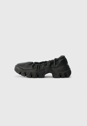 Juodų slip-on batų su raukšlėta odos viršutine dalimi ir storu tvirtu guminiu padu su giliais protektoriais, rodoma šoniniame profilyje ant balto fono.