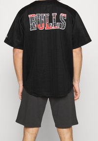 Jersey negro de manga corta con rayas verticales y "Bulls" en letras bold blancas y rojas en la parte posterior. Combinado con pantalones cortos grises.