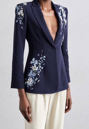 Marineblazer med blomsterbroderi og perledekorationer, båret med cremefarvede bukser, der viser en dyb V-hals og figursyet talje.