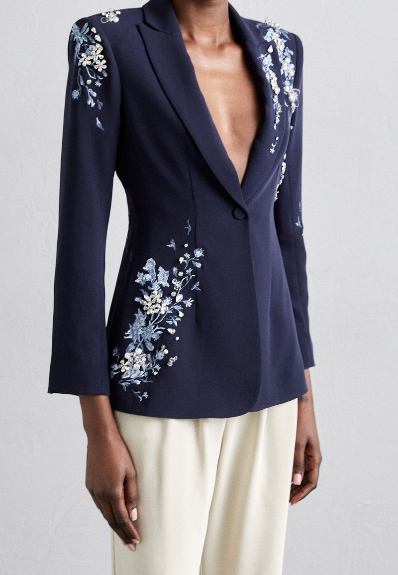 Blazer bleu marine avec broderie florale et accents en perles porté avec un pantalon crème, montrant un décolleté en V profond et une taille cintrée.