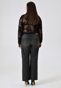 Zwarte doorschijnende blouse met bloemenpatroon, gecombineerd met hoge donkergrijze wijde broek. Zwarte hakken, gezien van achteren.