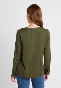 Blouse verte olive à manches longues avec un col rond, une texture lisse et des détails de boutons aux poignets. Portée avec un jean bleu.
