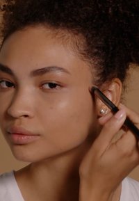 Application de maquillage avec un pinceau noir sur la joue. La personne a un teint lisse, brun clair et porte de petites boucles d'oreilles décoratives.