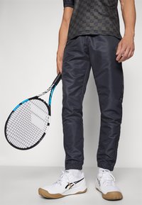 Pantalones deportivos de color gris oscuro con puños elásticos, combinados con zapatillas de tenis blancas y color beige. Se sostiene una raqueta de tenis con un acento azul en la mano derecha.