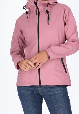 Personne portant une veste à capuche rose avec une fermeture éclair noire et des cordons, associée à un jean bleu, mains jointes devant.