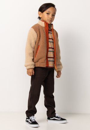 Junges Kind steht, trägt eine braune und beige Fleecejacke über einem gestreiften Shirt, dunklen Hosen und schwarz-weißen Turnschuhen vor einem weißen Hintergrund.