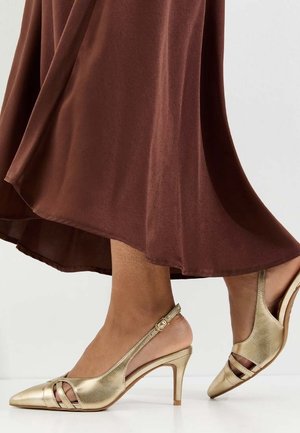 Pieds portant des chaussures à talons hauts dorées à bride arrière, avec une jupe marron fluide visible à partir du milieu du mollet vers le bas.