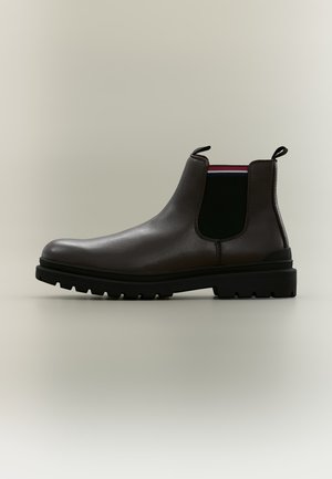 Botte Chelsea en cuir marron foncé avec panneaux latéraux élastiques noirs, languettes, et semelle épaisse à crampons noirs sur fond beige clair.