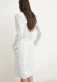 Robe florale blanche avec des motifs bleus, manches longues, longueur au genou, taille ceinturée et texture de tissu lisse. La vue de dos révèle un design simple.