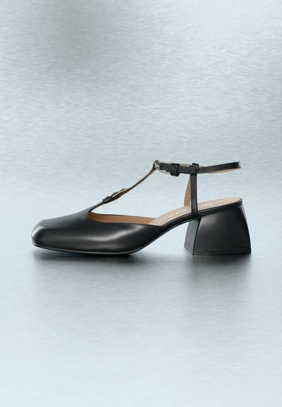 Μαύρο δερμάτινο παπούτσι με χαμηλό τετράγωνο τακούνι, λουράκι Τ και κούμπωμα σε γκρι αντανακλαστική επιφάνεια.