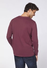 Langarm-Shirt in Kastanienrot aus leichtem Stoff, Rundhalsausschnitt, weiche Textur und gerader Saum, kombiniert mit dunkelblauen Jeans.