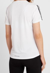 Camiseta atlética blanca con mangas cortas, rayas de acento negras en los hombros, tejido texturizado y un sutil detalle plateado en el dobladillo.