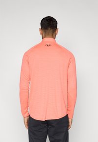 Κοραλλί αθλητική μπλούζα με μακριά μανίκια, υφασμένη υφή και ψηλό γιακά. Διαθέτει λογότυπο στο πίσω μέρος και κυρτή τελική ραφή.