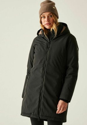 Kobieta w czarnej zimowej kurtce z kapturem i brązowej dzianinowej czapce, stojąca na tle jednolitego jasnego tła.