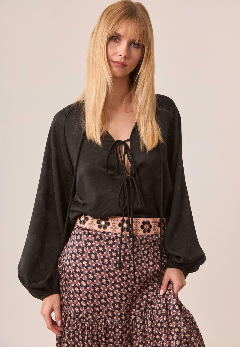 Blusa negra con mangas abullonadas y un detalle de lazo en el cuello, combinada con una falda rosa con estampado floral y un dobladillo con volantes.