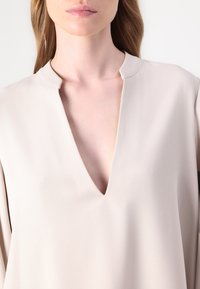Blusa beige con scollo a V profondo e maniche corte. Il tessuto ha una texture liscia e leggermente lucida con un design minimalista.