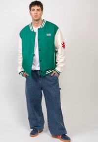 Zielono-biała kurtka varsity z prążkowanym kołnierzem i mankietami, z czerwonym motywem graficznym na rękawie. Luźne spodnie dżinsowe i niebieskie buty.