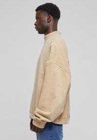 Beige oversized genser med høy krage, laget av mykt stoff. Har senkede skuldre og ribbede mansjetter, kombinert med blå jeans.