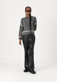 Grau gestrickter Pullover mit weißem geometrischen Muster, kombiniert mit schwarzen Hosen aus Kunstleder und schwarzen Schuhen. Das Model trägt Sonnenbrillen.