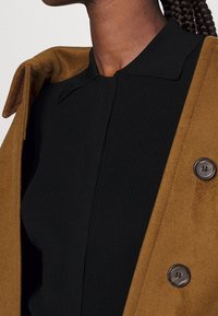 Brun ullkappa med krage, med knäppning framtill. Under finns en svart ribbad topp som tillför textur och djup.