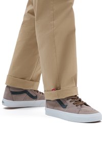 Sneakers in suede marrone con dettagli in verde scuro e suole bianche. Indossati con pantaloni beige a risvolto. Dettagli di cucitura intricati visibili.