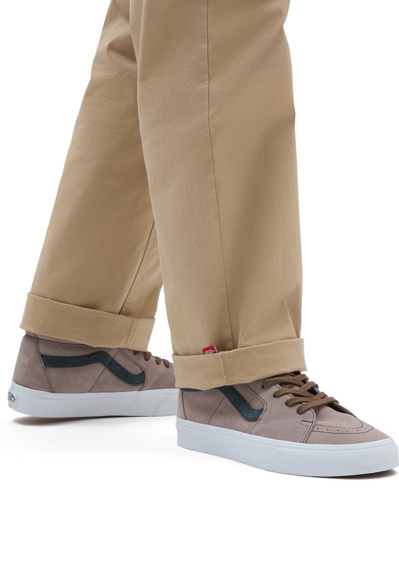 Sneakers in suede marrone con dettagli in verde scuro e suole bianche. Indossati con pantaloni beige a risvolto. Dettagli di cucitura intricati visibili.