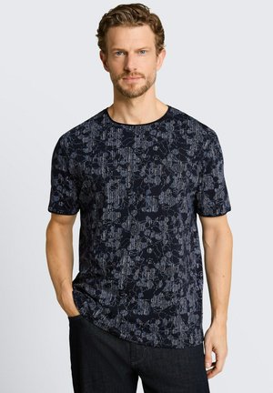 Mężczyzna z krótkimi kręconymi włosami, ubrany w ciemnoniebieską koszulę z krótkim rękawem w wzory i czarne spodnie, stoi z jedną ręką w kieszeni.