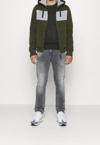 Uomo che indossa un zip-up felpa verde e grigia, una maglietta verde scuro, jeans grigi sbiaditi e sneaker bianche, in piedi su uno sfondo bianco.