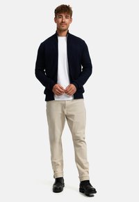 Marine bomberjakke med herringbone tekstur, hvit glidelås, båret over en hvit t-skjorte. Lette beige bukser og svarte sko fullfører antrekket.