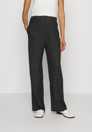 Person trägt dunkelgraue, weite Hose, weißes ärmelloses Oberteil und weiße, strukturierte Loafers, steht mit einer Hand in der Tasche.