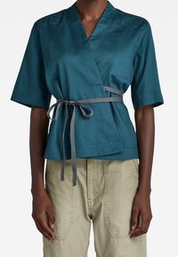 Personne portant un haut court à manches courtes de style cache-cœur bleu sarcelle avec ceinture grise à la taille et pantalon beige clair, debout devant un fond blanc.