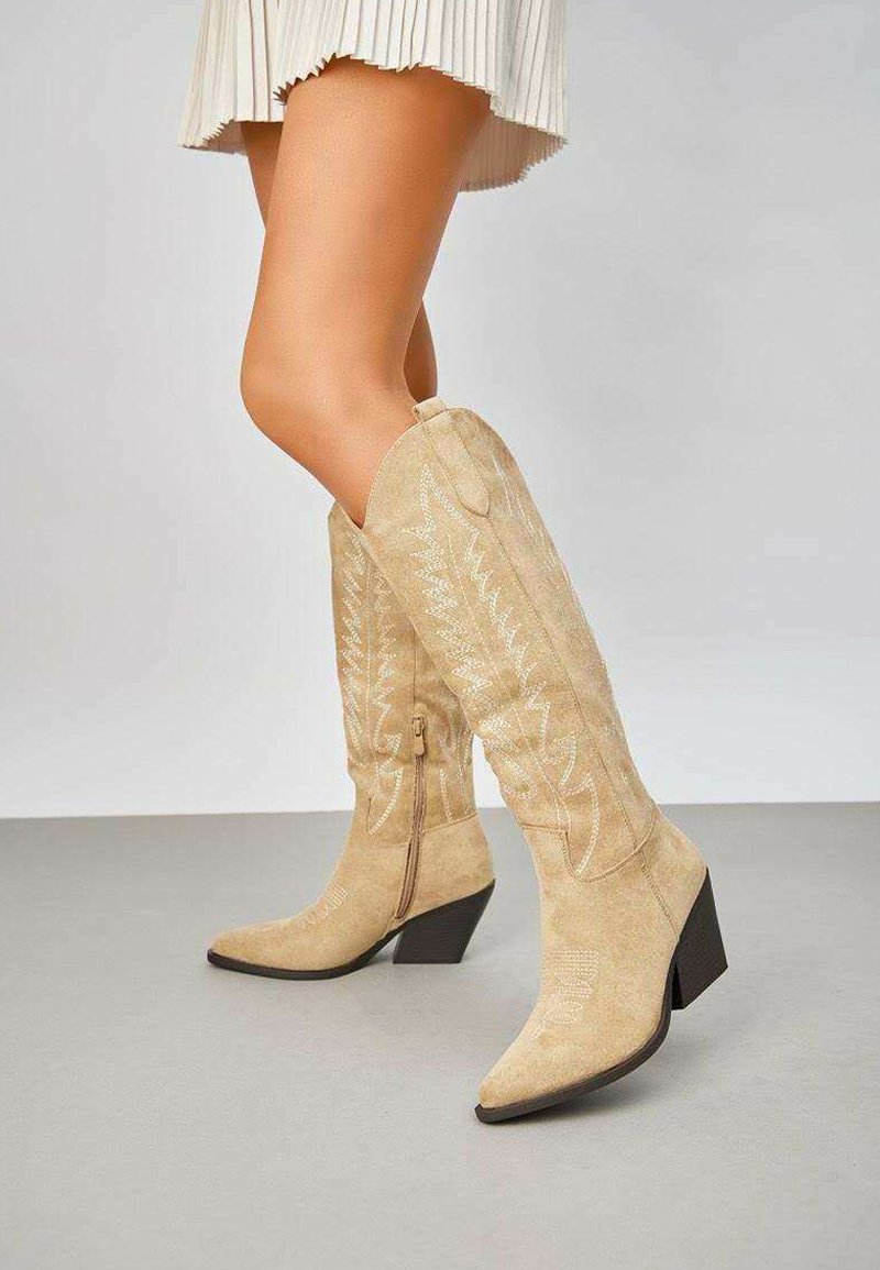 Bottes hautes en suède beige avec des pointes en forme de aiguille et des talons carrés. Motifs de couture décoratifs le long de la tige, avec une fermeture éclair sur le côté.