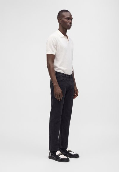 Homme debout portant un polo blanc à manches courtes, un jean noir et des mocassins noir et blanc devant un fond clair uni.