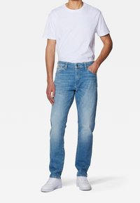 Hombre de pie vestido con una camiseta blanca sencilla, jeans azul claros y zapatillas blancas, con una mano en el bolsillo frente a un fondo blanco.